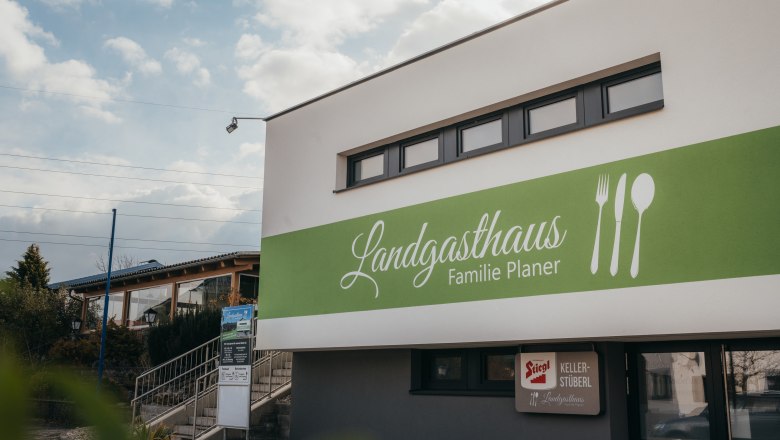 Außenansicht des Landgasthauses Familie Planer mit grünem Schild und Bestecksymbolen.