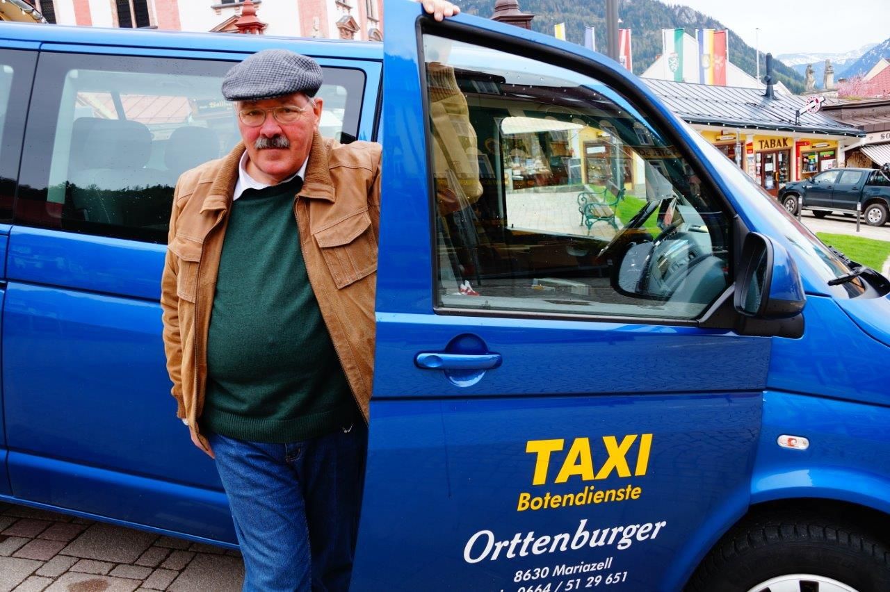 Ein Mann steht neben einem blauen Taxi mit der Aufschrift 'Taxi Botendienste Orttenburger'.