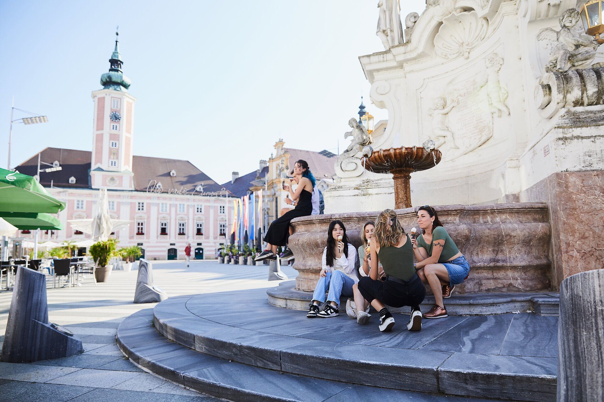 An einem sonnigen Tag versammeln sich fröhliche Menschen um einen prächtigen Brunnen, der die Atmosphäre des Rathausplatzes belebt. Die historische Architektur im Hintergrund und die bunten Fahnen verleihen dem Ort einen festlichen Charakter, während die frische Luft und das Lachen der Freunde eine einladende Stimmung schaffen.