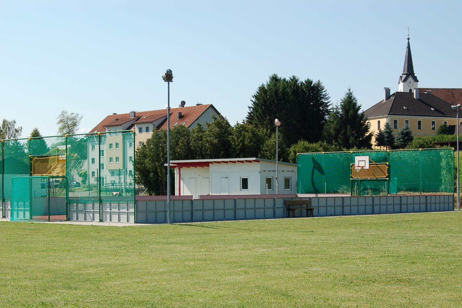 Ein Funcourt mit Basketballkorb und grünem Netz, daneben ein kleines Gebäude. Im Hintergrund sind Wohnhäuser und eine Kirche zu sehen.