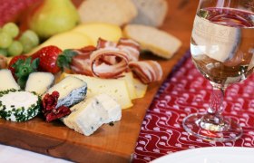 Eine Brettljause mit Käse, Schinken, Brot und Obst auf einem Holzbrett, daneben ein Glas Weißwein.