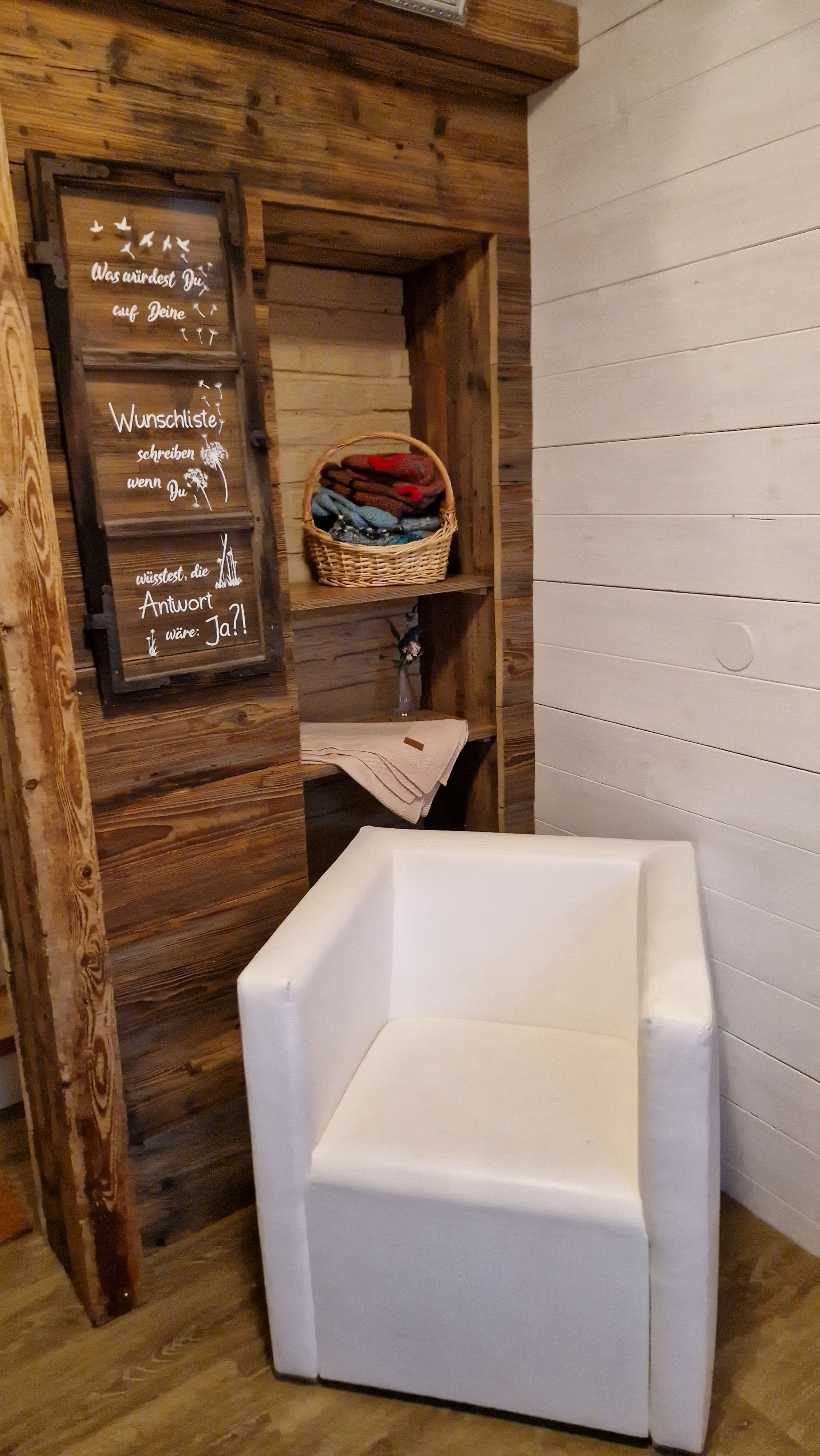 Ein gemütlicher Raum mit einem weißen Sessel, einem Regal mit gefalteten Decken und einer Tafel mit inspirierendem Text.