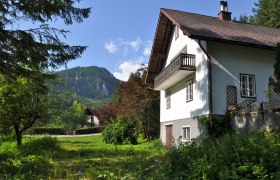 Ein wei&szlig;es Ferienhaus mit Balkon vor einer Berglandschaft und gr&uuml;nem Garten.