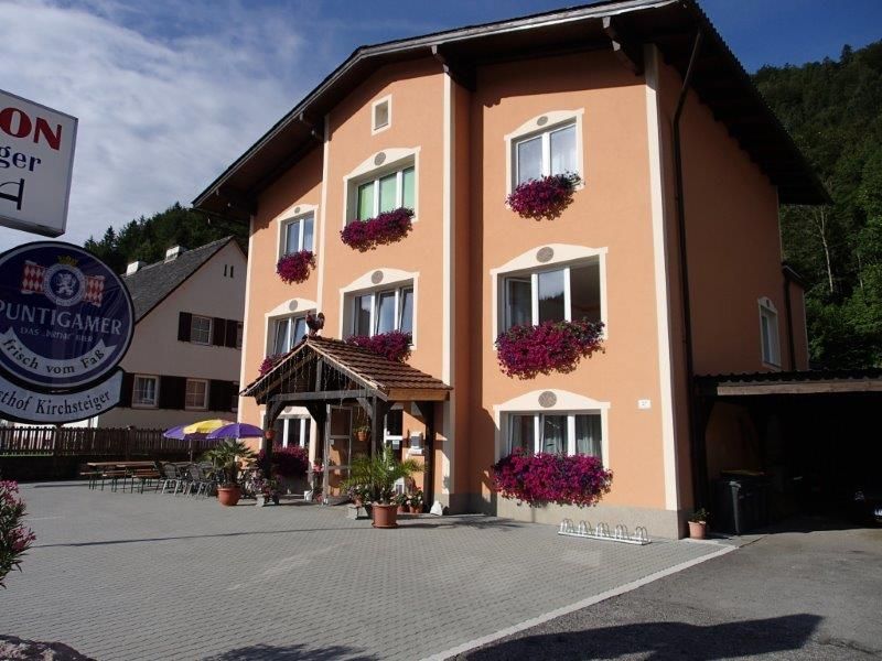 Ein dreistöckiges Gebäude mit orangefarbener Fassade und Blumenkästen vor den Fenstern. Ein Schild mit der Aufschrift 'Puntigamer' ist sichtbar.