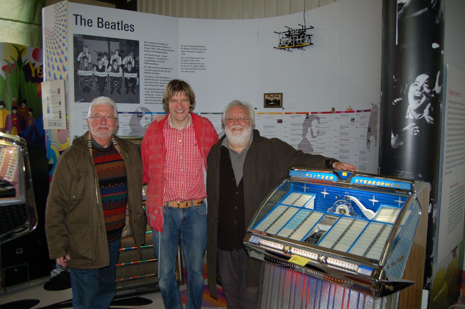 Drei Männer stehen vor einer Beatles-Ausstellung mit einer Jukebox.