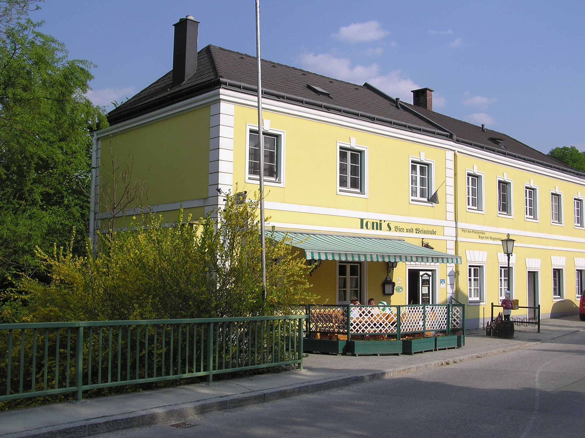 Gelbes Gebäude mit der Aufschrift 'Toni's Bier- und Weinstube', umgeben von grünen Pflanzen und einem Zaun.