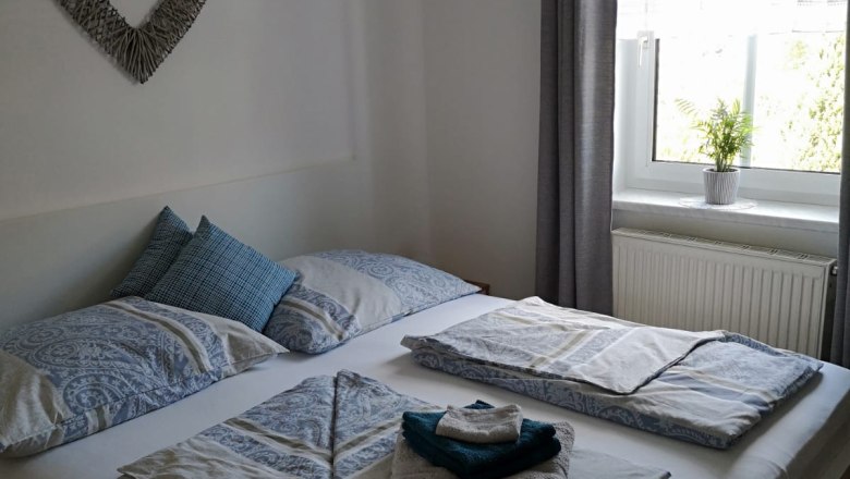 A cozy bedroom with a double bed, decorated with blue bed linen and towels. A picture of a heart hangs on the wall, a window with a curtain next to it.