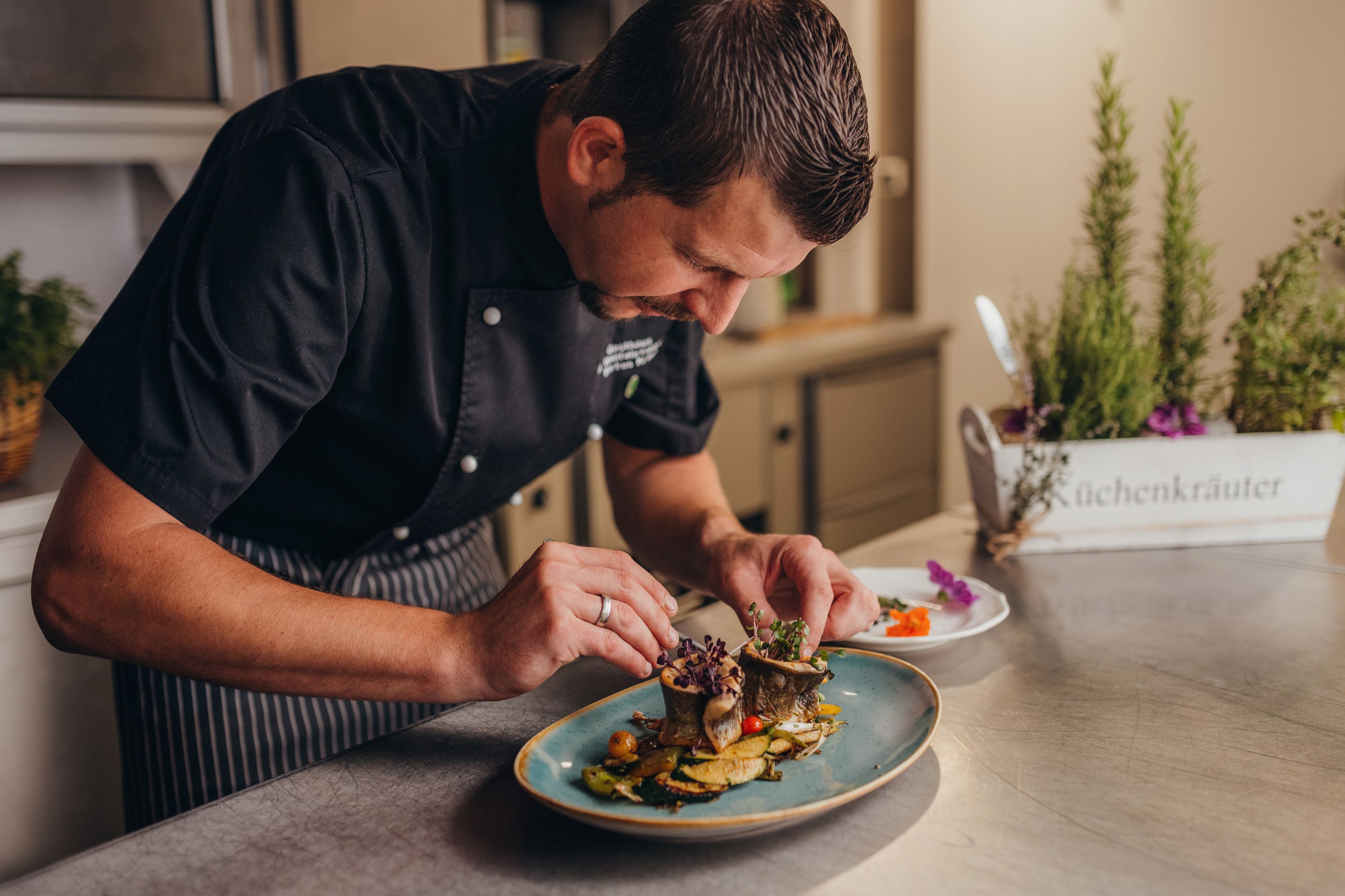 Ein Koch richtet ein Gericht auf einem Teller an, im Hintergrund sind Küchenkräuter zu sehen.