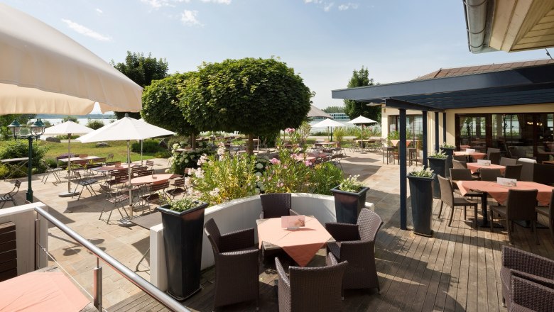 Sonnige Terrasse mit Tischen, Stühlen und Sonnenschirmen, umgeben von Pflanzen und Bäumen.