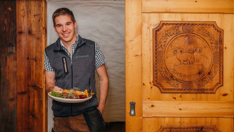 A man in traditional dress holds a plate of food in front of a wooden door with a carved motif.