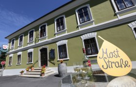 Grünes Gebäude mit weißen Fenstern, davor ein Tisch mit Weinflasche und Gläsern, Schild mit Aufschrift 'Most Straße'.