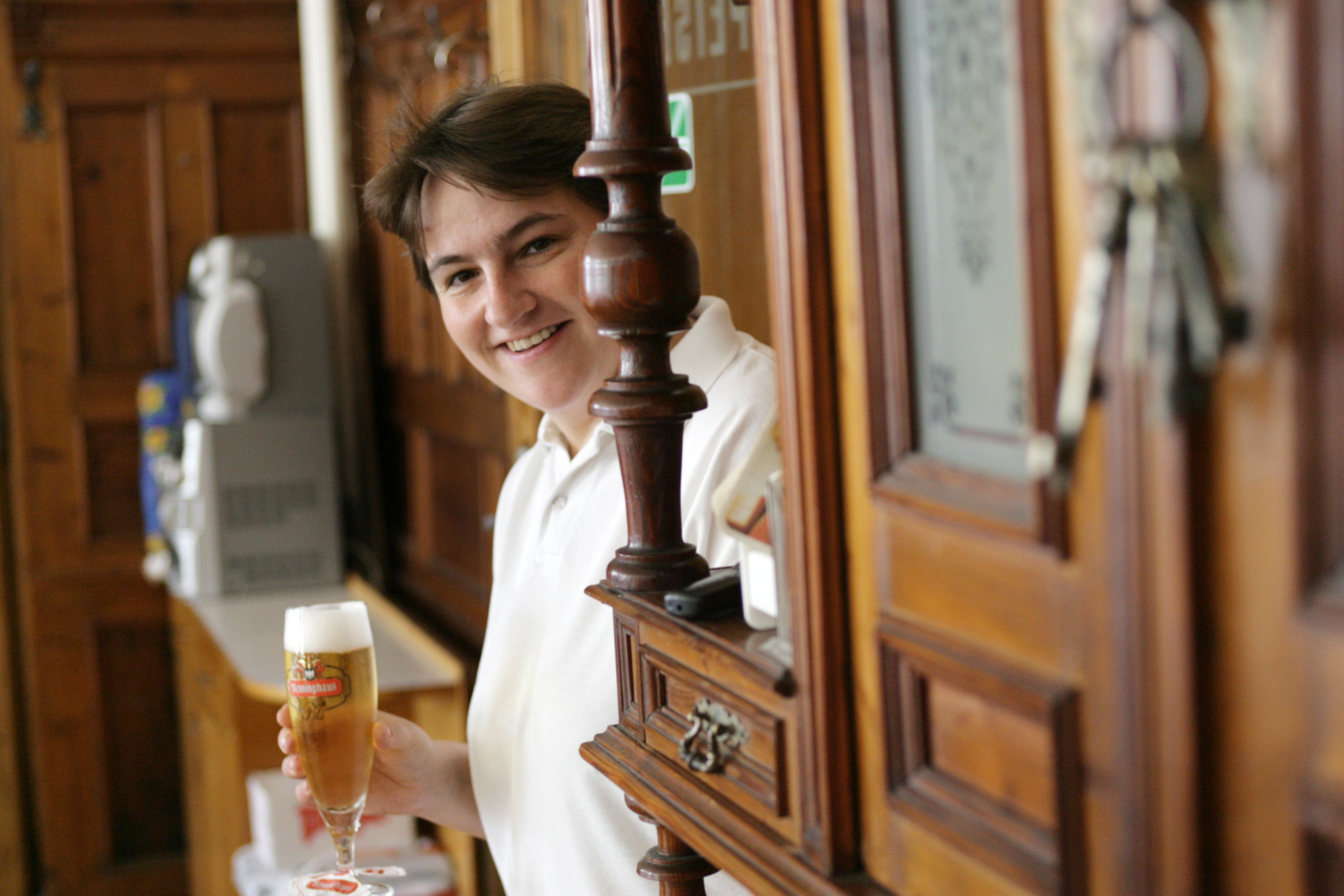Person in einem Gasthaus mit einem Glas Bier in der Hand, lächelnd hinter einer Holzbar.