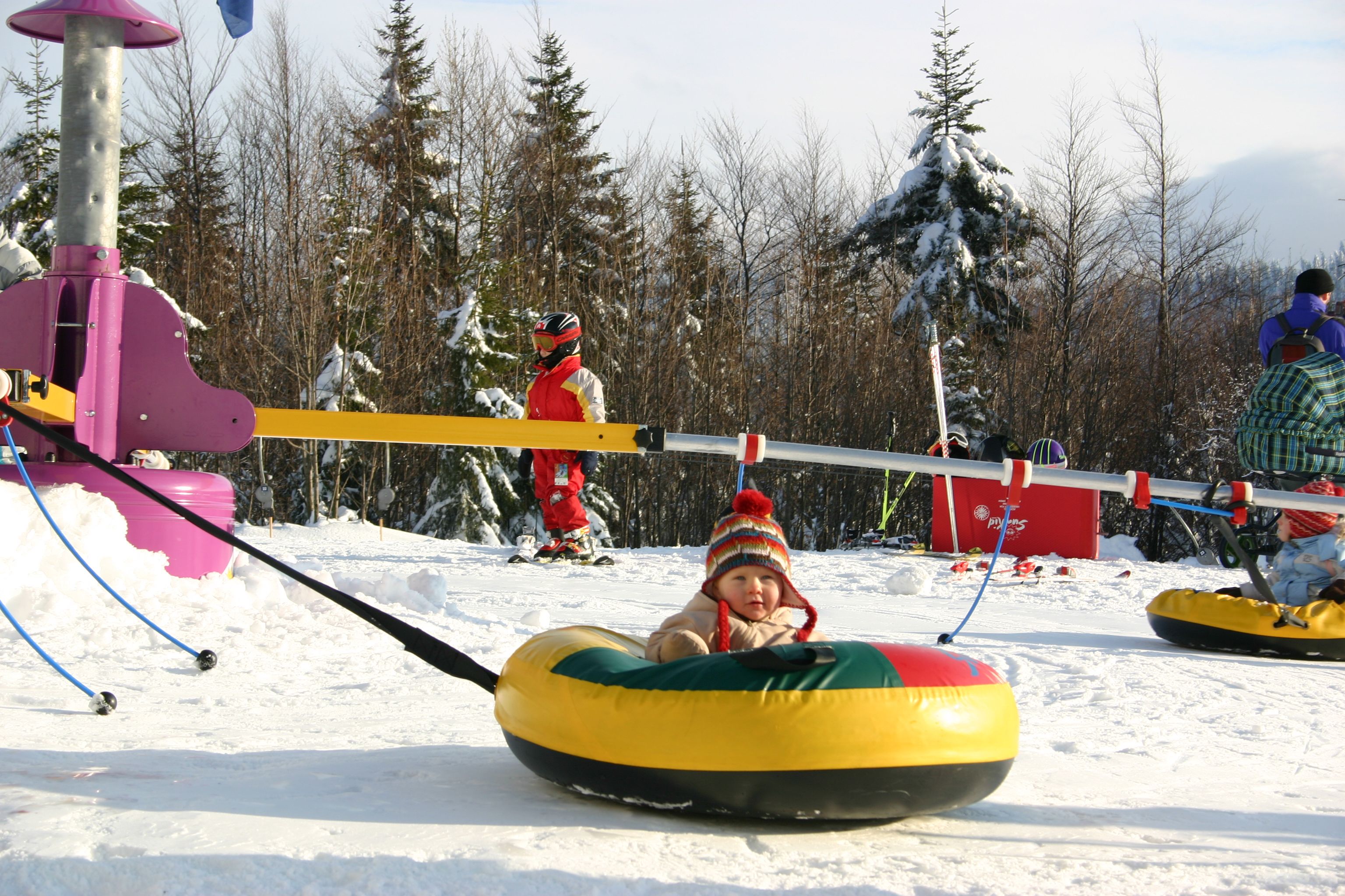 Kleinkind in einem bunten Schneereifen auf einem verschneiten Hügel mit anderen Kindern im Hintergrund.