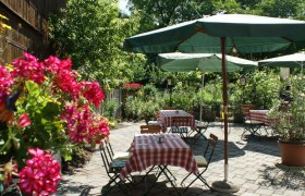 Ein gem&uuml;tlicher Gastgarten mit rot-wei&szlig; karierten Tischdecken, gr&uuml;nen Sonnenschirmen und bl&uuml;henden Blumen im Vordergrund.
