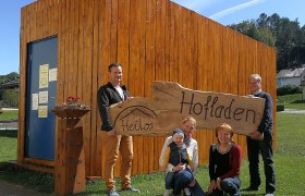 Familie vor einem Holzgebäude mit einem Schild 'Heilos Hofladen'.