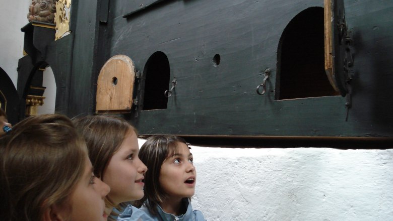 Three children look curiously at a wall with small doors.