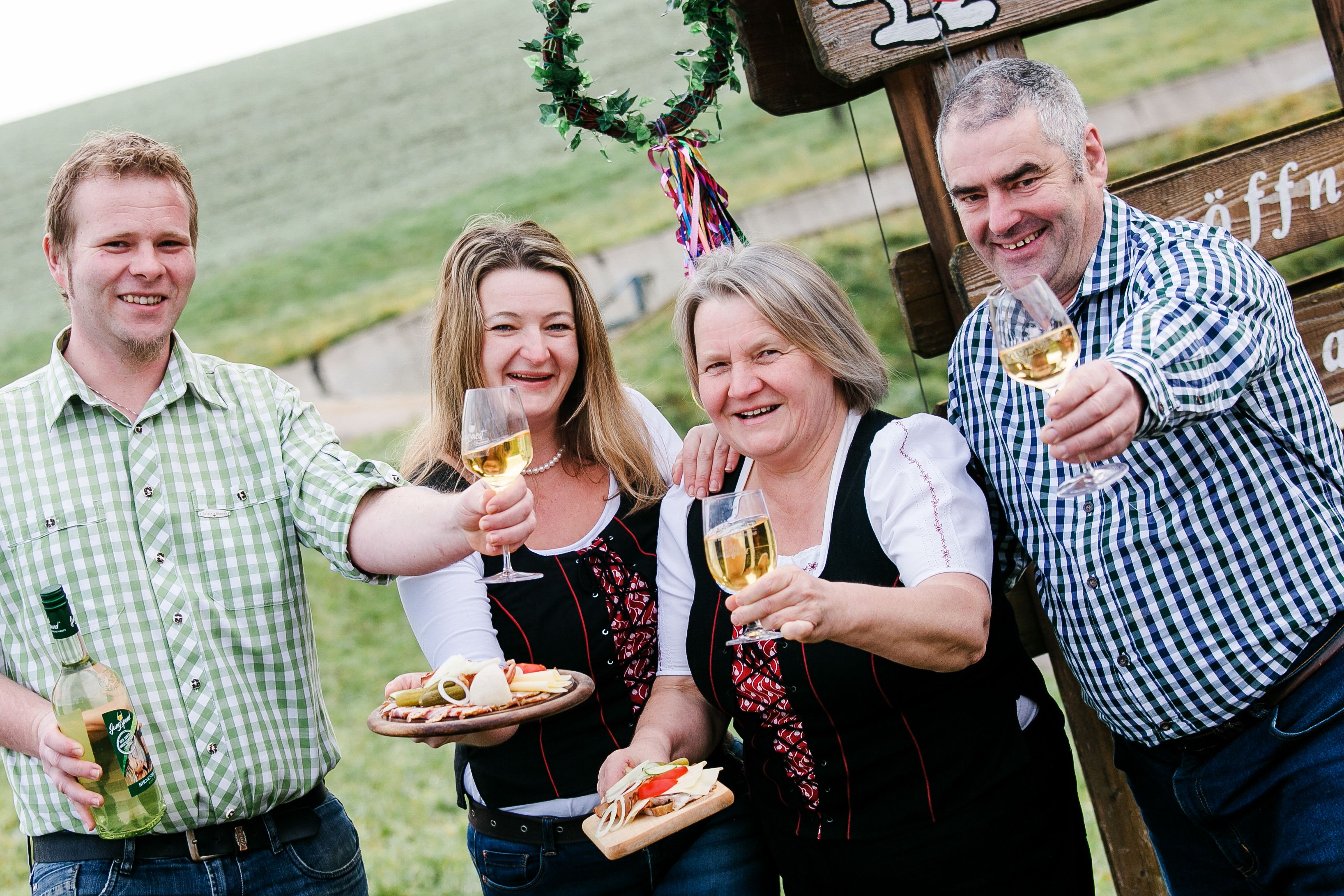 Vier Personen stoßen mit Weingläsern an, im Freien vor einem Schild.