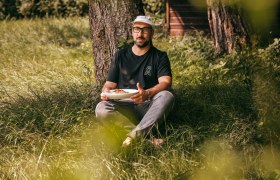 Mann sitzt im Gras unter einem Baum und hält einen Teller mit Essen.