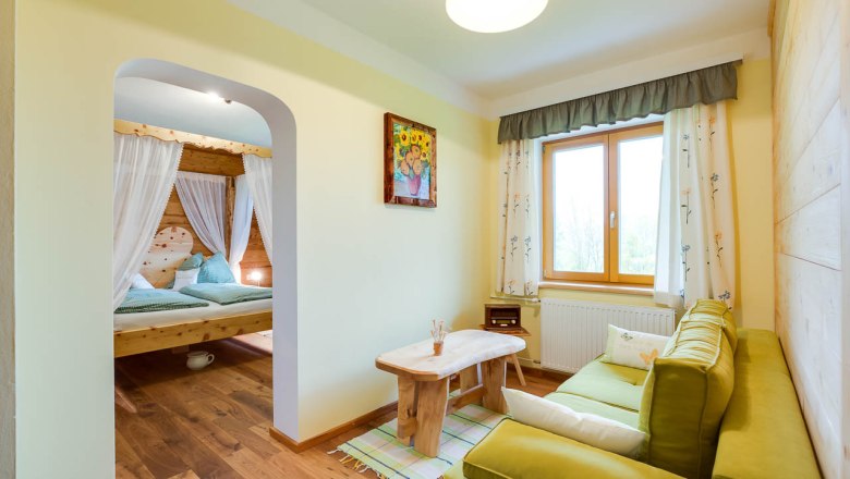 Gemütliches Zimmer mit Holzmöbeln, grünem Sofa, großem Fenster und einem Himmelbett im Hintergrund.