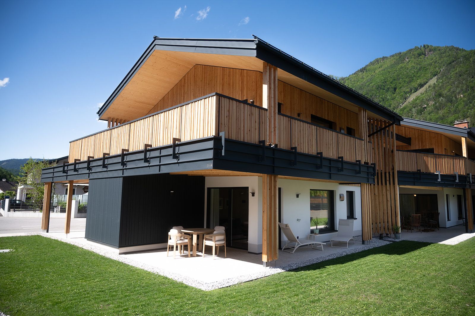 Moderne Holzarchitektur eines zweistöckigen Gebäudes mit Balkon und Terrasse, umgeben von grüner Landschaft.