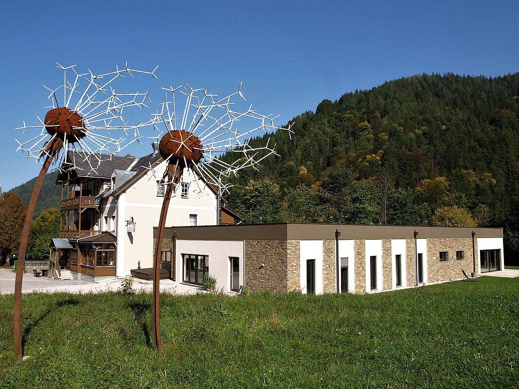 Ein modernes Gebäude mit großen Löwenzahn-Skulpturen im Vordergrund, umgeben von grünen Hügeln und Bäumen.