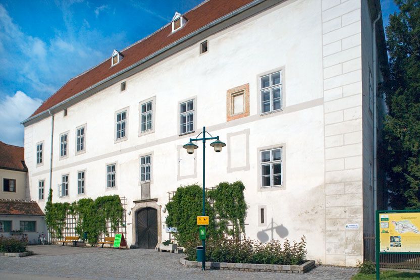 Schloss Traismauer mit weißer Fassade und roten Dachziegeln, umgeben von Pflanzen und einem gepflasterten Vorplatz.