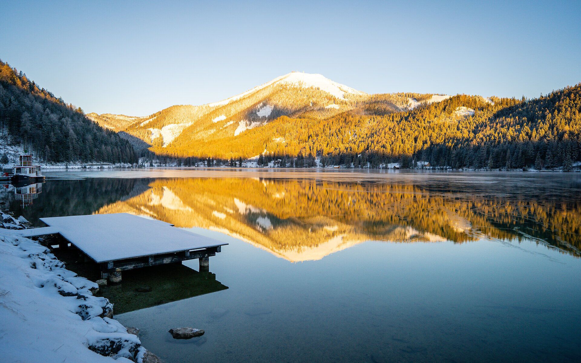 Die sanften Hügel sind mit einer glitzernden Schneedecke bedeckt, während die Sonne die Bergspitzen in warmes Gold taucht. Der ruhige See spiegelt die majestätische Landschaft wider und lädt zu einem besinnlichen Spaziergang ein. Hier, in der unberührten Natur, entfaltet sich die winterliche Pracht in voller Schönheit.