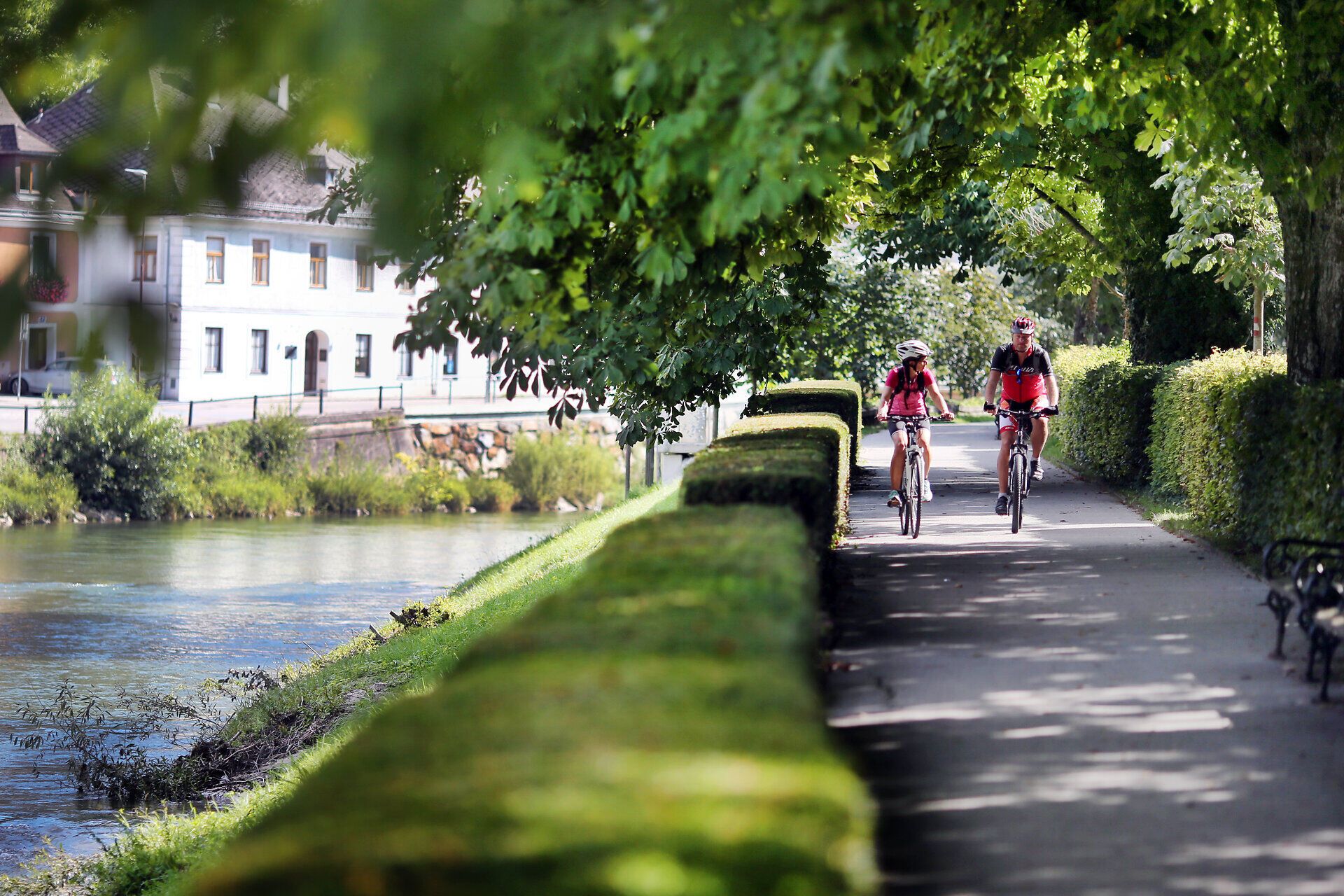 Ein Paar radelt entspannt entlang des malerischen Erlauftalradwegs, umgeben von üppigem Grün und dem sanften Plätschern des Wassers. Die schattenspendenden Bäume und die idyllische Landschaft laden dazu ein, die frische Luft und die Schönheit der Natur zu genießen.
