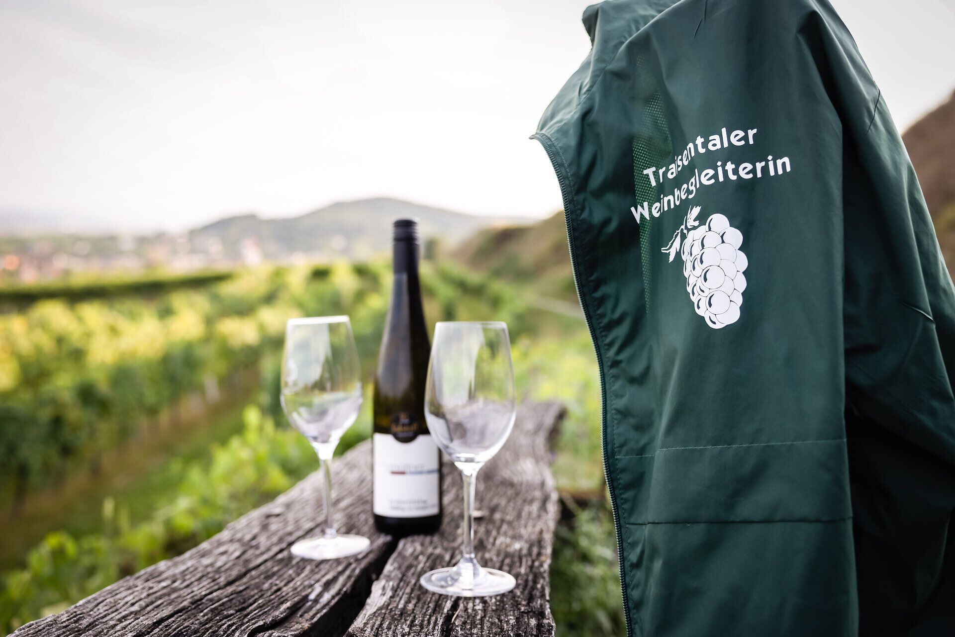 Auf einem alten Holzbalken stehen zwei Weingläser und eine Flasche Weißwein, daneben hängt eine Jacke mit den Traisentaler Weinbegleitern