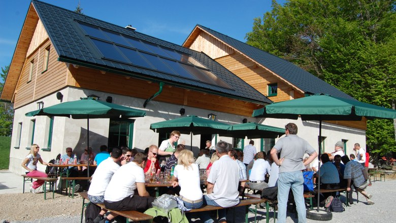 Menschen sitzen an Tischen vor einer H&uuml;tte mit Solarpanelen und Sonnenschirmen.