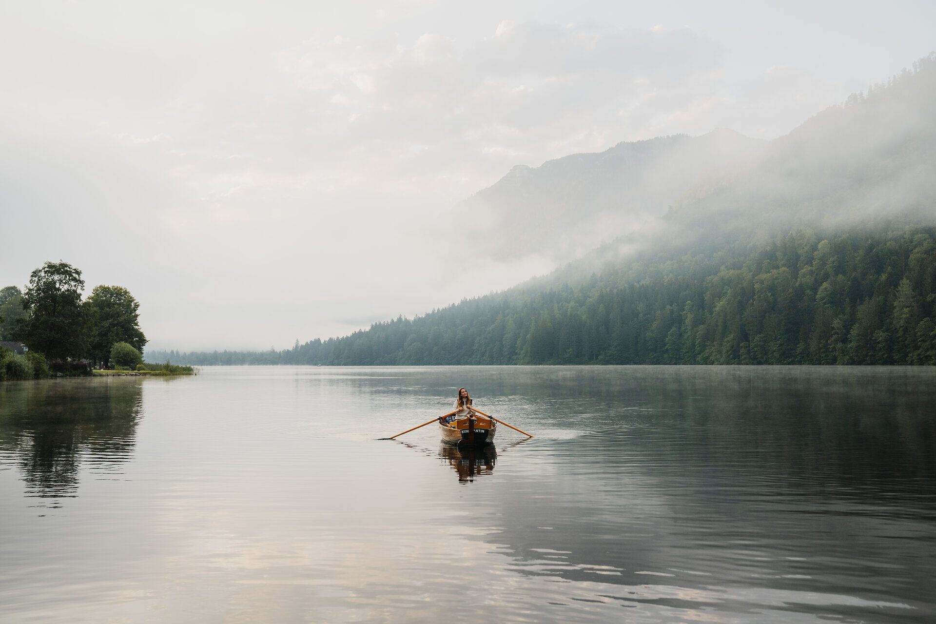 Sanfte Nebelschwaden umhüllen den ruhigen See, während ein Boot gemächlich über die spiegelglatte Wasseroberfläche gleitet. Die umliegenden Berge, majestätisch und grün, laden zu unvergesslichen Erlebnissen in der Natur ein.