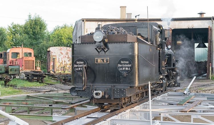 Dampflokomotive vor einem Heizhaus mit Schienen im Vordergrund.