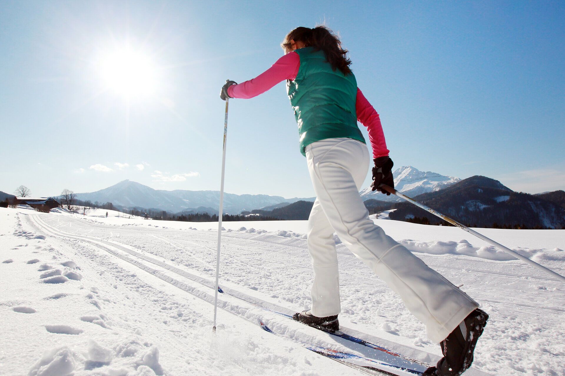 In der glitzernden Winterlandschaft gleitet eine Frau elegant über die schneebedeckten Loipen. Die strahlende Sonne taucht die Umgebung in ein warmes Licht und schafft eine einladende Atmosphäre für Langlaufbegeisterte. Umgeben von majestätischen Bergen und der frischen, klaren Luft ist dies der perfekte Ort, um die winterliche Stille zu genießen.