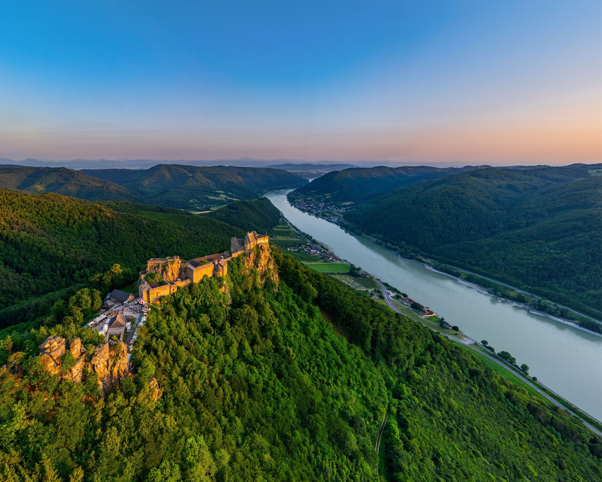 Luftaufnahme der Burgruine Aggstein an der Donau, umgeben von grünen Hügeln und Wäldern bei Sonnenuntergang.