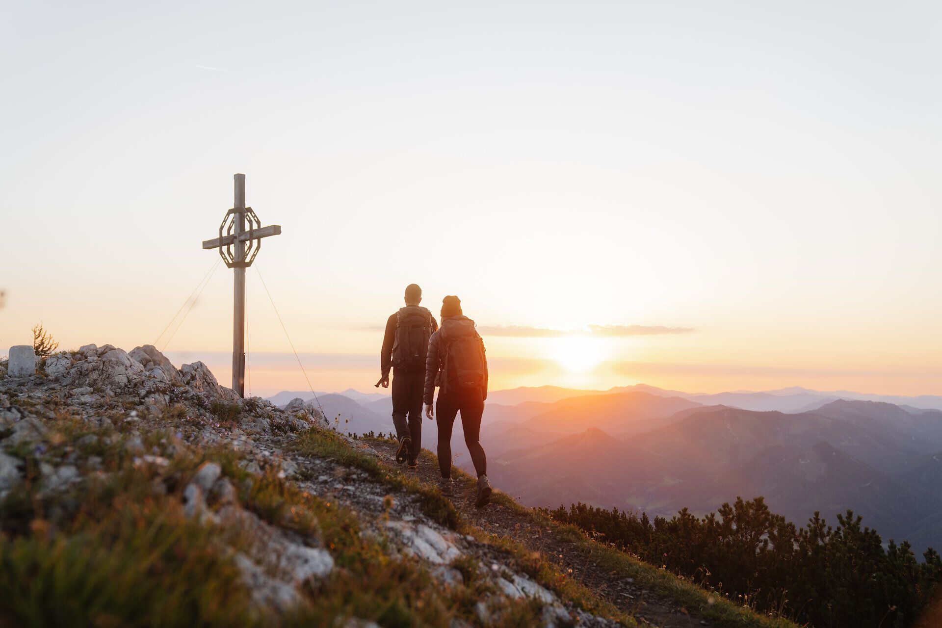 Ein Paar wandert Hand in Hand den malerischen Pfad entlang, während die Sonne hinter den sanften Hügeln des Mostviertels untergeht. Das Gipfelkreuz strahlt in der Abenddämmerung und lädt dazu ein, die atemberaubende Aussicht auf die umliegenden Berge zu genießen. Hier, wo die Natur in voller Pracht erblüht, wird jeder Schritt zu einem unvergesslichen Erlebnis.