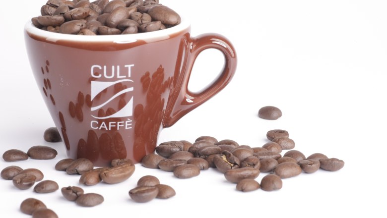 Braune Tasse mit Aufschrift 'Cult Caffè', gefüllt mit Kaffeebohnen, auf weißem Hintergrund.