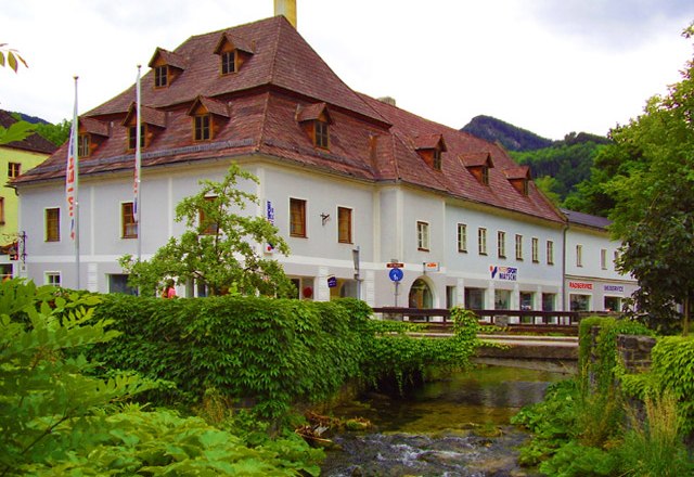 Historisches Gebäude mit rotem Dach an einem Bach, umgeben von grüner Vegetation.