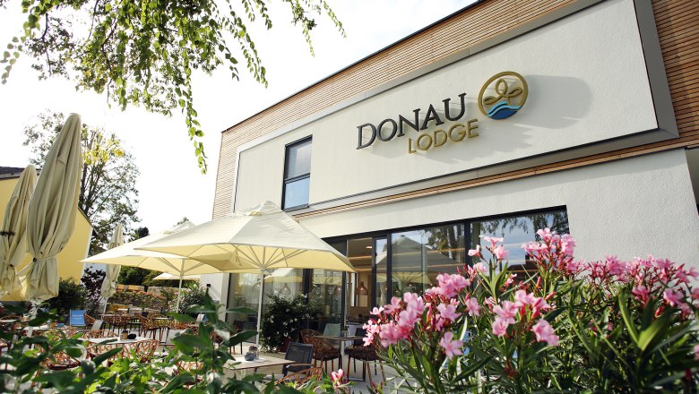 Außenansicht der Donaulodge mit Terrasse, Sonnenschirmen und blühenden Pflanzen.