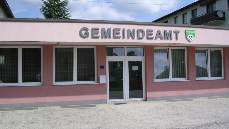 Au&szlig;enansicht eines rosa Geb&auml;udes mit der Aufschrift 'GEMEINDEAMT' und einem Wappen.