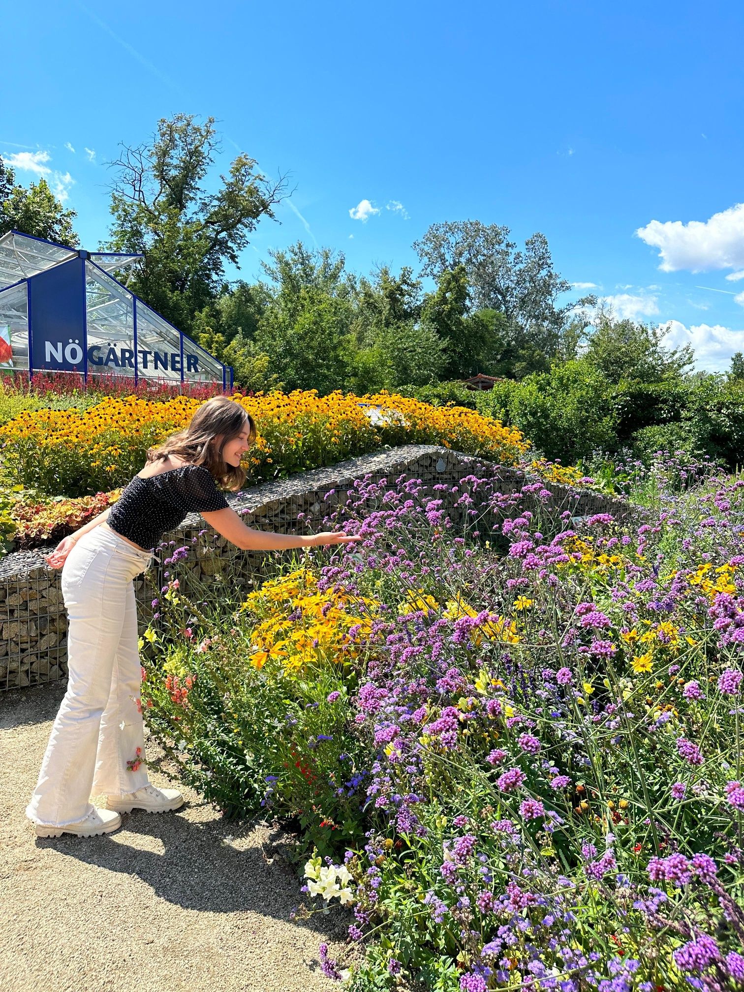 Eine Frau beugt sich über ein Blumenbeet in einem Garten mit einem Gewächshaus im Hintergrund.