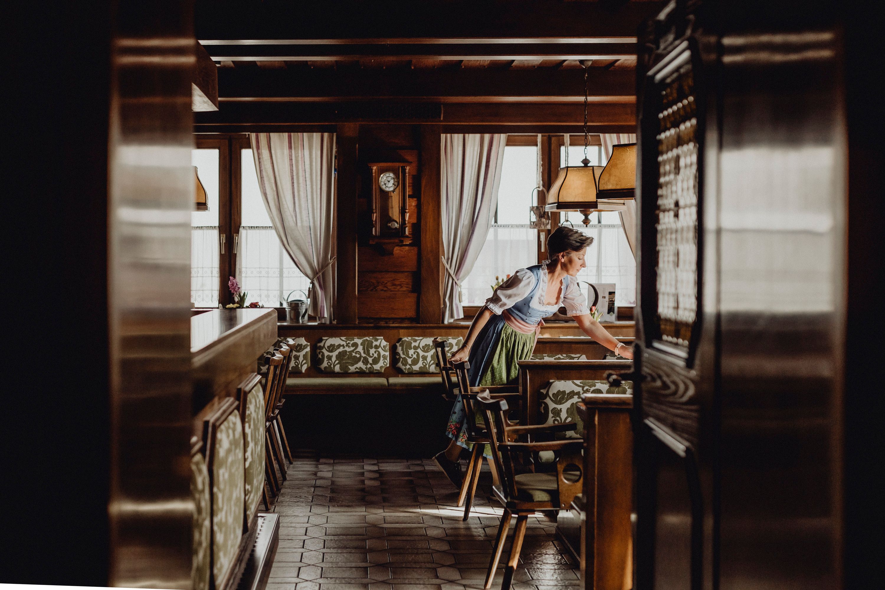 Eine Frau in traditioneller Kleidung richtet einen Tisch in einem gemütlichen, holzgetäfelten Gastraum her.