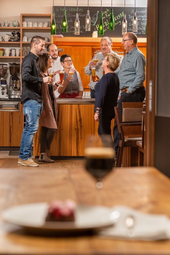 Eine Gruppe von Menschen steht in einer Bar und hält Biergläser in der Hand. Im Vordergrund sind ein Glas und ein Dessert auf einem Tisch zu sehen.