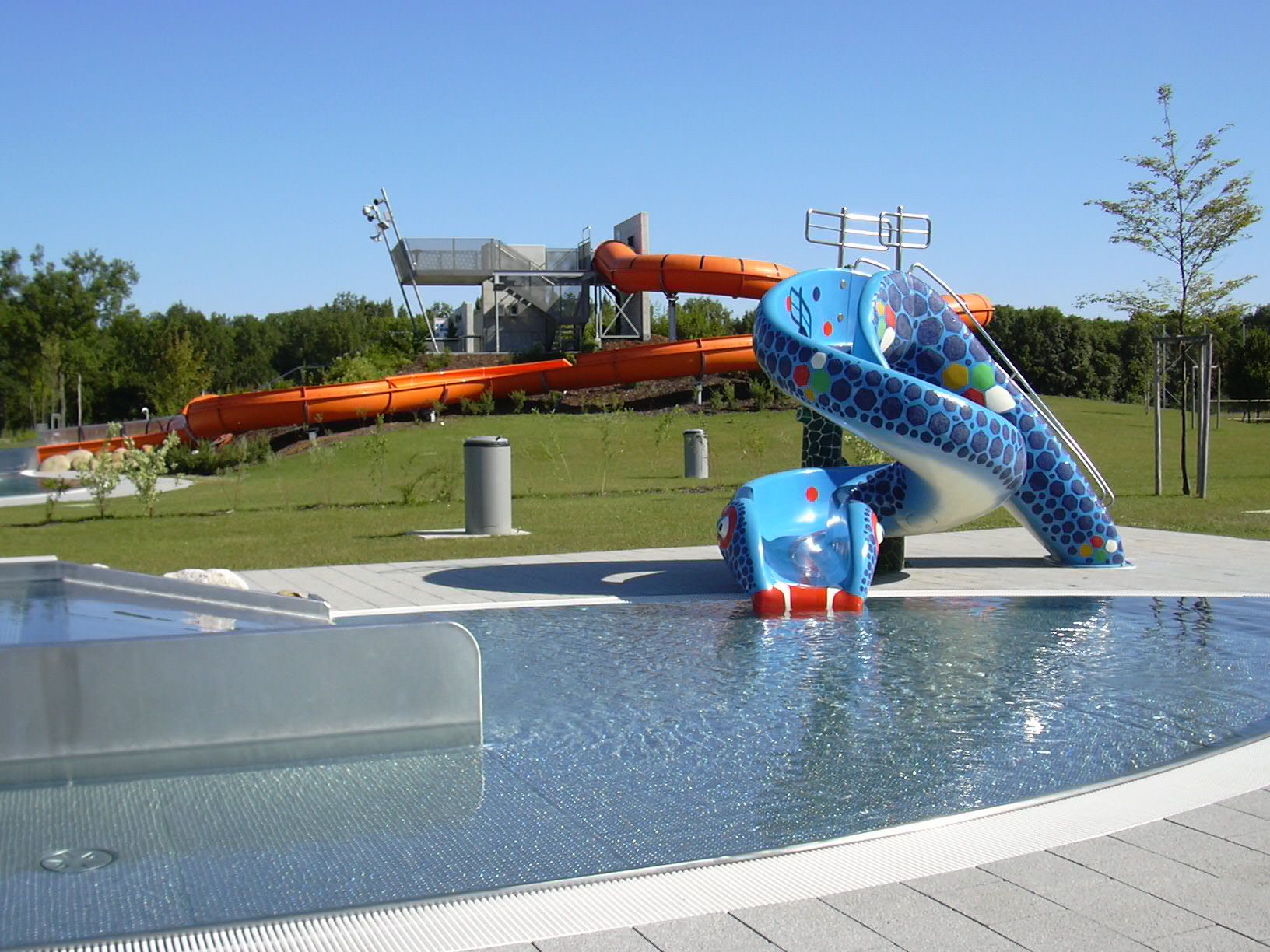 Blick auf einen Aquapark mit blauer Kinderrutsche und großer oranger Wasserrutsche im Hintergrund.