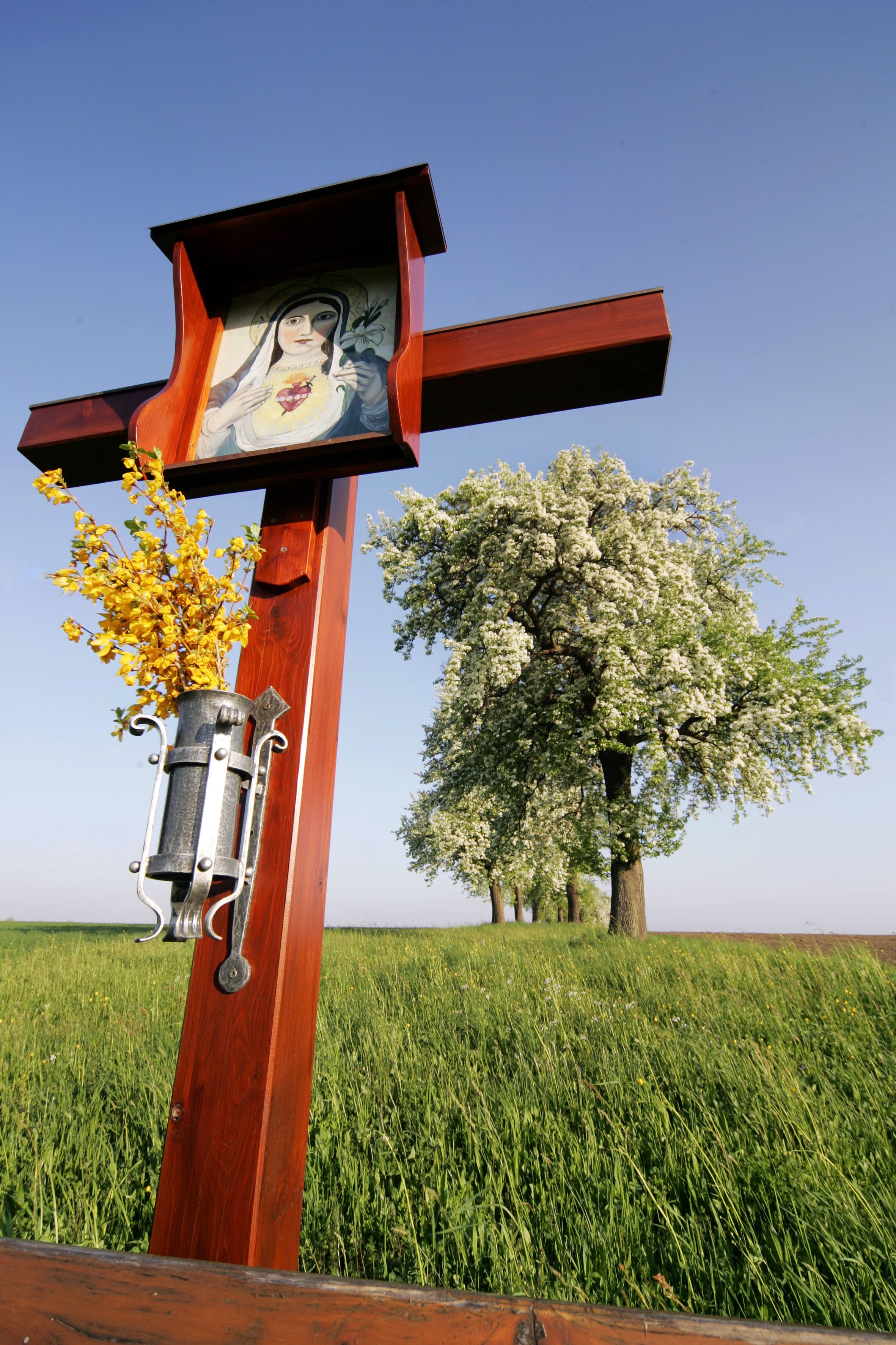 Ein hölzernes Wegkreuz mit einem Bild der Jungfrau Maria und gelben Blumen, im Hintergrund blühende Bäume auf einer Wiese.