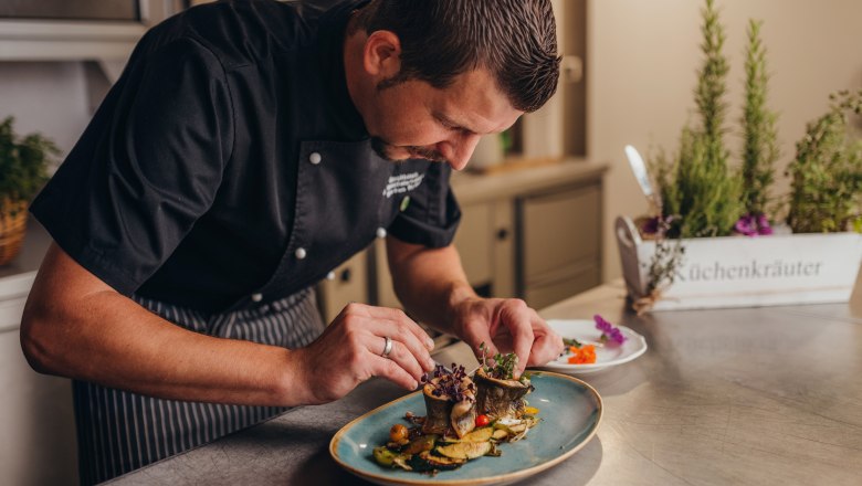 Ein Koch richtet ein Gericht auf einem Teller an, im Hintergrund sind Küchenkräuter zu sehen.