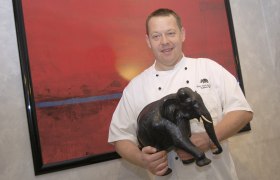 Ein Koch in weißer Uniform hält eine schwarze Elefantenfigur vor einem roten Gemälde.