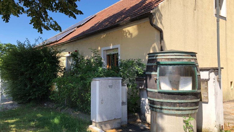 A yellow house with a red tiled roof and solar panels. In the foreground, a large barrel with a sight glass and plants.