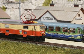 Modelleisenbahn mit Lokomotive und Waggon vor einem Miniaturbahnhof.