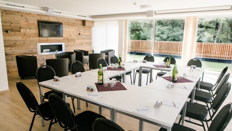 Konferenzraum mit U-förmiger Tischanordnung, Stühlen, Holzwand, Fernseher und großen Fenstern mit Blick ins Grüne.