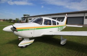 Ein kleines Flugzeug, Piper Cherokee Archer II, steht auf einer grünen Wiese vor einem Hangar und einem Kontrollturm.
